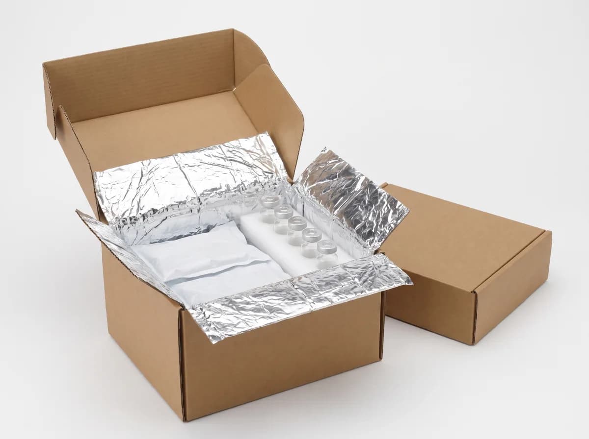 Open insulated shipping box lined with reflective thermal foil, holding two flat gel cold packs and a row of lyophilised vials nested in white foam